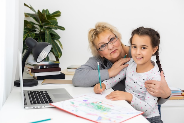 Mature grandmother helping child with homework at home. Satisfied old grandma helping her granddaughter studying in living room. Little girl writing on notebook with senior teacher sitting next to her