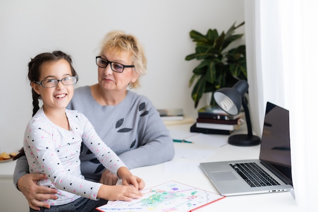 Mature grandmother helping child with homework at home. Satisfied old grandma helping her granddaughter studying in living room. Little girl writing on notebook with senior teacher sitting next to her