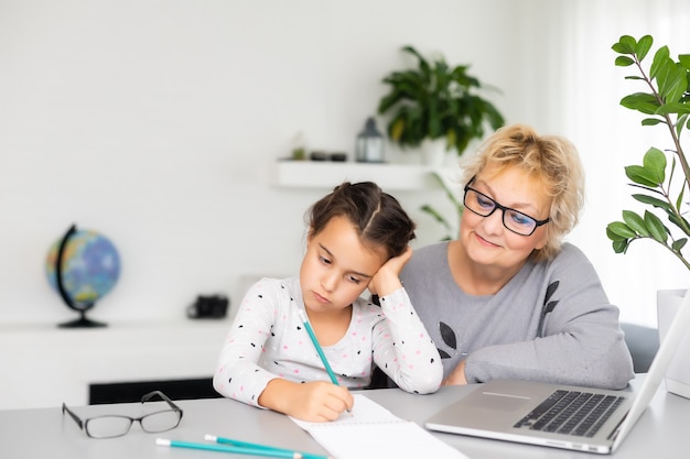 Mature grandmother helping child with homework at home. Satisfied old grandma helping her granddaughter studying in living room. Little girl writing on notebook with senior teacher sitting next to her