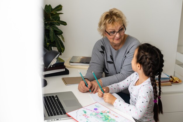 Mature grandmother helping child with homework at home. Satisfied old grandma helping her granddaughter studying in living room. Little girl writing on notebook with senior teacher sitting next to her