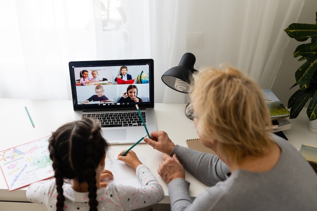 Mature grandmother helping child with homework at home. Satisfied old grandma helping her granddaughter studying in living room. Little girl writing on notebook with senior teacher sitting next to her