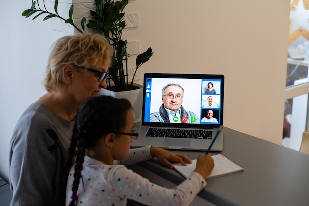 Mature grandmother helping child with homework at home. Satisfied old grandma helping her granddaughter studying in living room. Little girl writing on notebook with senior teacher sitting next to her