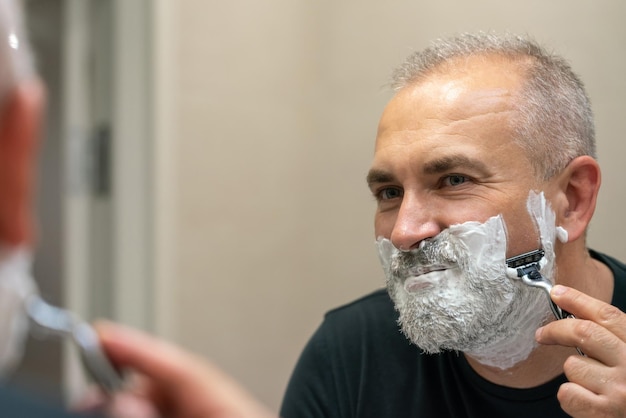 Mature grayhaired man restyling his beard himself at home using razor