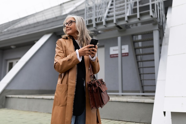 Mature grayhaired stylish business woman rushing to a meeting\
with a mobile phone in her hands