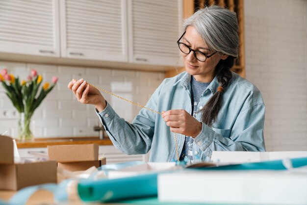 Mature grey woman in eyeglasses wrapping present at home