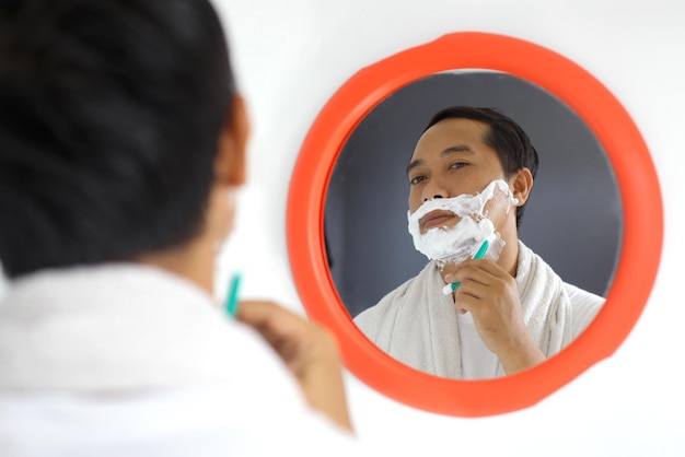 Mature handsome Asian man shaving in front of mirror