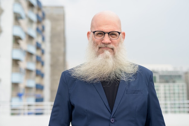 Mature handsome bald bearded businessman wearing eyeglasses against view of the city