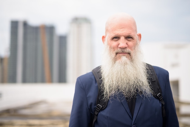 Photo mature handsome bald bearded businessman with backpack against view of the city