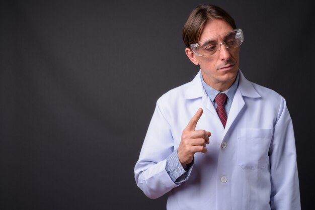 mature handsome Italian man doctor wearing protective glasses against gray wall