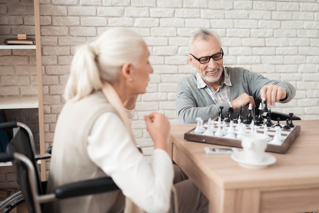 Mature happy couple in wheelchairs play chess.