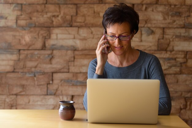 Mature happy woman smiling while talking on mobile phone and using laptop on wooden table against brick wall