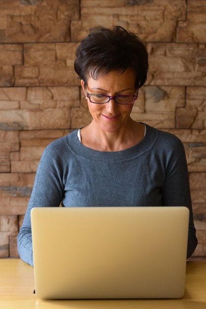 Mature happy woman smiling while wearing eyeglasses and using laptop on wooden table against brick wall