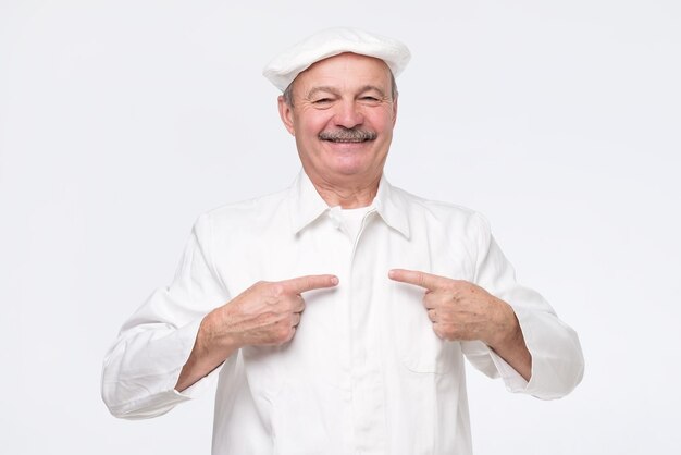 Mature hispanic chef in white uniform pointing to self being proud