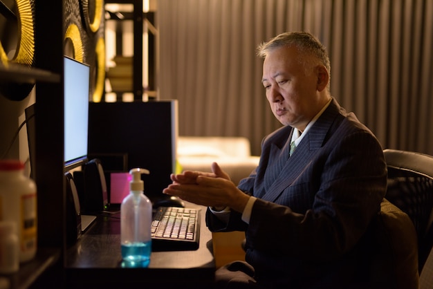 Mature Japanese businessman using hand sanitizer while working overtime at home late at night