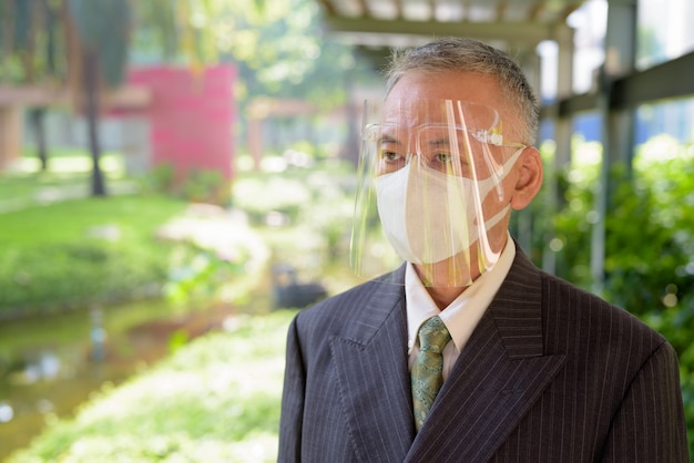 Mature Japanese businessman with mask and face shield thinking at the park