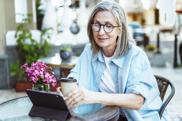 Photo mature lady enjoying outdoors while searching for information on tablet pc woman seated at cafe table on street sipping cup of coffee moment of leisure and relaxation