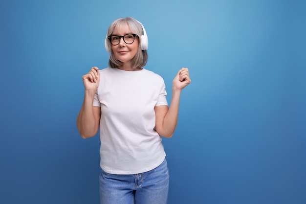 Mature lady with gray hair listens to music in wireless headphones on studio background with copy