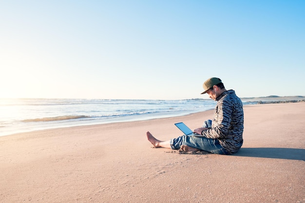 Зрелый латиноамериканец сидит на песке пляжа со своим ноутбуком и работает на закате