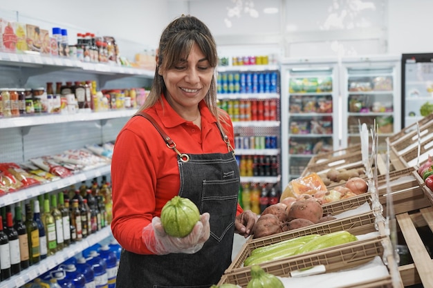 Mature latin woman working inside supermarket while holding\
fresh oragnic zucchini