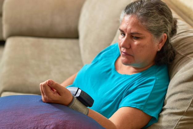 Mature Latina woman measures her blood pressure with digital monitor from home Hypertension Concept