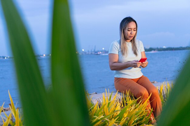 Mature Latina woman sitting on the dock over the lake, using a smart phone.