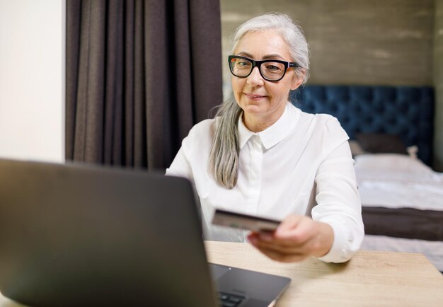 Mature long haired woman wearing glasses sitting with laptop and buy something at the internet in her comfortable flat