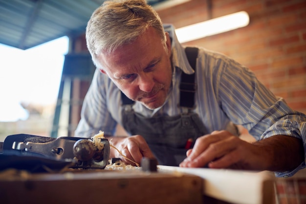 Mature Male Carpenter In Garage Workshop Planing Piece Of Wood