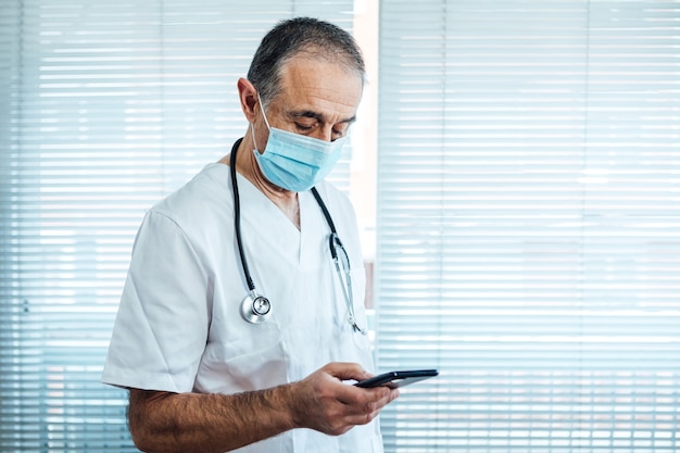 Mature male doctor - nurse wearing face mask, looking at mobile phone next to a hospital window. Covid-19 and medicine concept