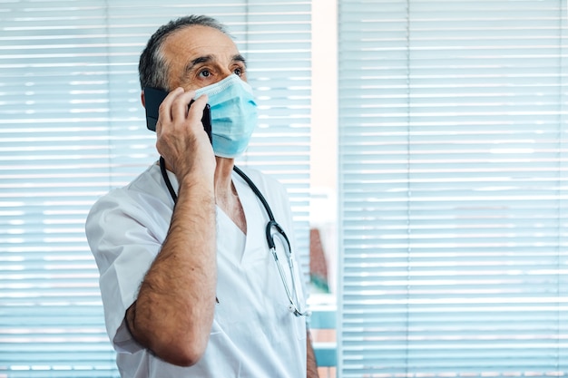 Mature male doctor - nurse wearing face mask, talking on mobile phone next to a hospital window. Covid-19 and medicine concept