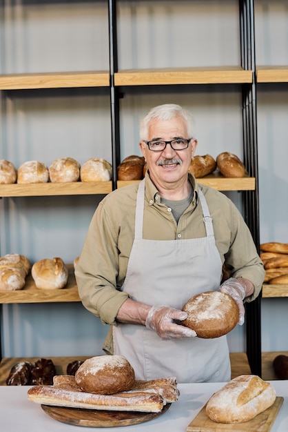 Mature man in apron and eyeglasses holding loaf of fresh bread