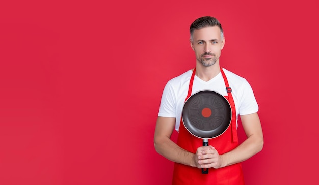 Mature man chef in apron hold frying pan on red background