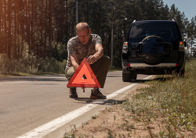 Mature man driver putting triangle caution and warning road sign on asphalt near car after accident ...