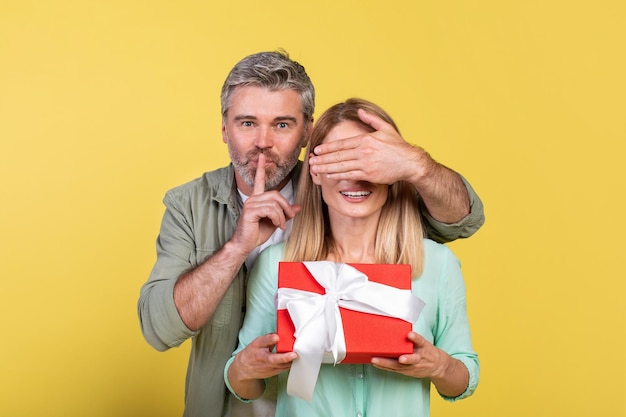 Photo mature man got surprise for his loving wife woman holding gift box man covering her eyes and showing silence gesture