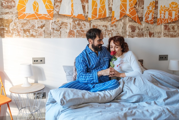 Mature man on his pajamas giving a rose on valentines day to her wife relaxed at home in bed

