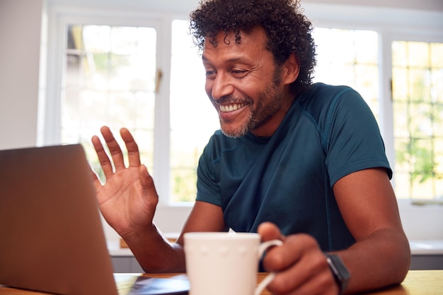 Mature Man At Home In Kitchen Waving As He Makes Video Call On Laptop Computer