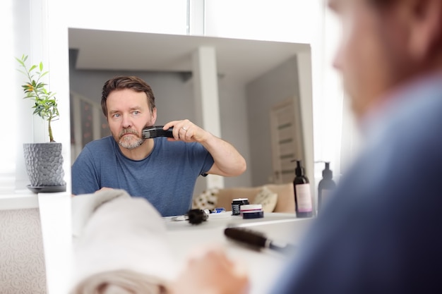 Photo mature man is shaving off his beard with electric razor at home during quarantine. handsome bearded man trimming his beard with a trimmer at home while barbershops closed. reflection in mirror.