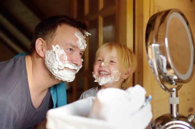 Mature man and little boy having fun with foam during shaving together. Kid son imitates his father.