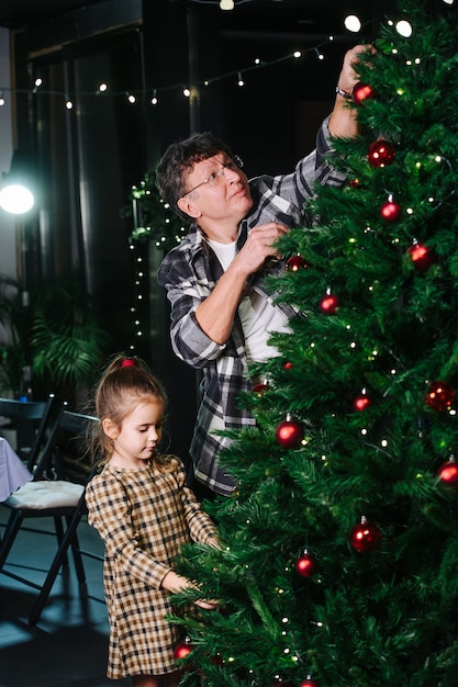 Mature man and little girl decorating a christmas tree with red shiny balls. Indoors