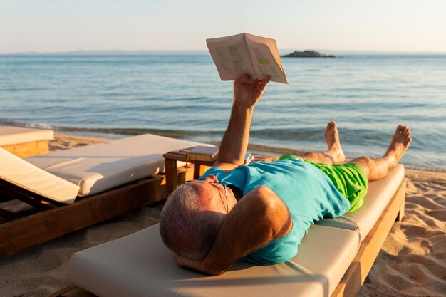 Mature man lying on a sunbed and reading a book at sunset summer vacation concept