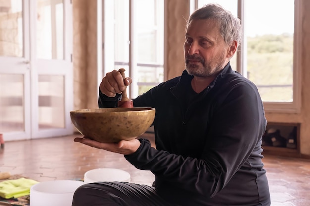 A mature man playing a bronze bowl seated and illuminated by light streaming through the window