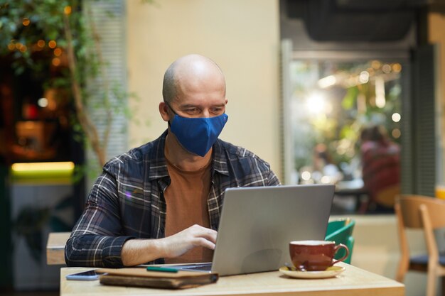 Photo mature man in protective mask working online on laptop while sitting in coffee shop