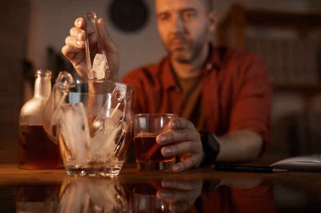 Uomo maturo che mette il ghiaccio nel bicchiere con alcool mentre è seduto al tavolo a casa