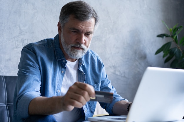 Mature man shopping online on laptop, using credit card at home.