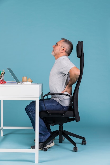 Uomo maturo che si siede nella sedia che soffre dal mal di schiena mentre usando sul computer portatile