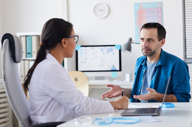 Mature man sitting opposite his doctor and telling about his complaints on the health at hospital