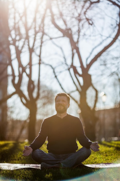 Uomo maturo seduto su materassino da yoga in comode asana sorridente e rilassante nel parco meditazione in condizioni di vita cittadina