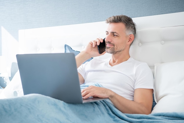 Mature man speaking on cellphone and working on laptop in bed