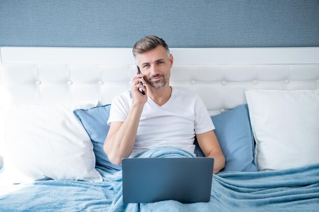 mature man speaking on phone and using laptop in bed