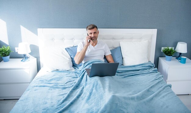 Зрелый мужчина разговаривает по телефону и работает за компьютером в постели