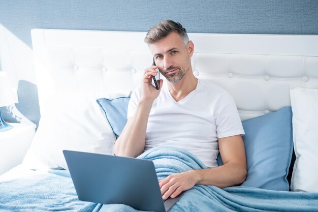 Mature man speaking on phone and working on laptop in bed freelance